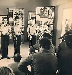 Three women holding sticks and wearing military uniforms and top-hats are performing to a crowd of soldiers. There are posters set up in the background. A shirtless male soldier is also in the background, on the right.