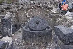 Circular basalt millstone set inside a matching basin in a stone-walled installation at Khirbet Wadi Hammam, with press weights and visitors walking past the ruins in the background