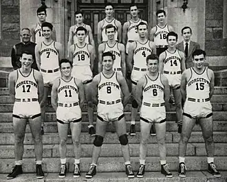 Three rows of five young men in white basketball uniforms stand on stone steps.