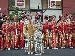 Girls in uniforms, headdresses, and white shoes stand waiting on the street. Two girls in the front face each other wearing pastel headdresses with feathers and multicolored uniforms with yellow trim. The girls in the back wear red uniforms with gold accents.