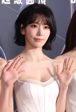 Sakura Miyawaki poses with her hand on her face, wearing a black blazer and decorative chain across her face.
