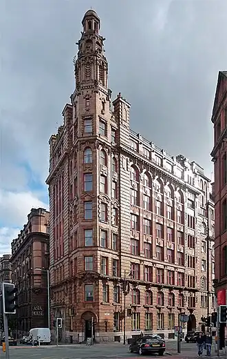 A red brick and terracotta building that sits on a corner of a square junction. The corner of the building is slightly rounded into an octagon-like shape with it then stretching above the building to become a clock tower.