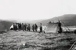A black and white photo of a group of Alaska Native men, women, and children alongside two dogs and one white explorer. They are standing in a small campsite with two tents overlooking a river valley