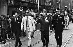 Photograph of the men in suits walking down a street with onlookers on each side. Panu Toivonen wear white. There are horse mounted patrolmen behind them on white horses.