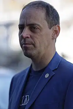 Portrait of a man with a short dark brown hair wearing a blue suit jacket and t-shirt.
