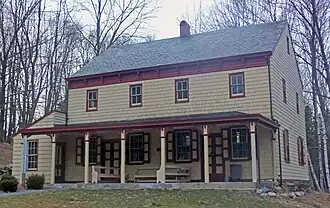 A two-story light yellow house with reddish trim, a pointed roof on the side, a small shed-roofed addition on the left and a porch along the front.