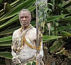 A man with white body paint covering his arms and torso holding a bow and arrows while smoking a cigarette