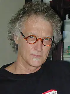 Portrait of a man with curly gray hair, round glasses, and a small patch of facial hair below his lower lip, wearing a black shirt indoors