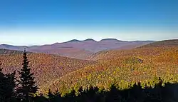 A landscape of forested mountains, in autumn color, underneath a blue sky, dominated by three rounded peaks in the distance with dark tops