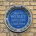 Round blue plaque with green floral surround and white text, reading "John Wesley (1703–1791) Lived here"
