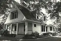 A black-and-white photo of a two-story house with a steep gabled roof, front porch, and large windows, partially shaded by surrounding trees