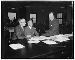 Black-and-white photo of a group of bankers in suits huddled together during a hearing recess.