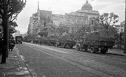 Trucks travelling along wide tree-lined street with large old building in the background