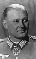 The head of a man, shown in semi-profile. He wears a military uniform with a military decoration in shape of an iron cross displayed at the front of his shirt collar. His hair is combed to back.