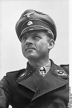 A man wearing a black military uniform, peaked cap and a neck order in the shape of a cross. His cap has an emblem in shape of a human skull and crossed bones.