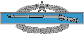 A metal device depicting a blue bar with a rifle, in front of a wreath of silver leaves.