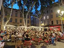 Crowded sidewalk café in Aix-en-Provence at dusk, with people dining outdoors under trees and surrounded by historic buildings.