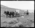 A man standing next to two horses that are harnessed to a wagon and another man is riding a horse and holding a spare horse behind the wagon. Taken outdoors.