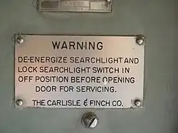 A metal plate attached to a piece of equipment with rivets. The plate reads "WARNING: De-energize searchlight and lock searchlight switch in off position before opening door for servicing / The Carlisle & Finch Co."
