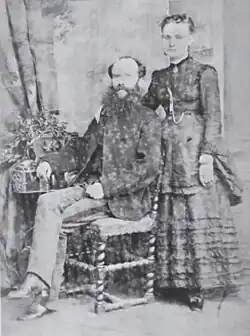 A bearded man sitting in a chair. A woman in a dress is standing behind him with her hand on his shoulder