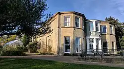 Building in natural honey coloured stone with three bays each having two storeys in a garden setting against a blue sky