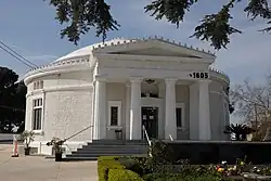 Photograph of the outside of a large circular building with pillars near the entrance. It has ‘1605’ written in black letters above and to the right of the entranceway.