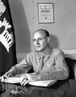 Man in shirt and tie with a West Point class ring sits at a desk. A telephone and writing pens are on the table. In the background is a US Army Corps of Engineers flag and a framed award from the National Safety Council.