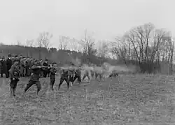 A large group of men standing in an open field, firing rifles during a live-fire exercise; smoke is visible around the shooters.