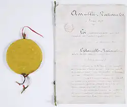 Yellow wax seal placed next to sheets of white paper bearing handwritten ink writing.