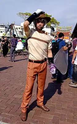 A man dressed as Joseph Joestar standing outdoors. He sports a grey-haired wig and a brown beard with a dyed grey moustache. He wears a fedora, a tan short-sleeved shirt with a brown stripe on the chest, orange-brown khaki pants, brown dress shoes, and white gloves. Wrapped around the man's right hand is a purple thorn-covered vine symbolizng Hermit Purple, Joseph's stand.