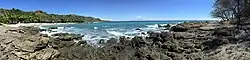 Panoramic view of a white sand beach and the turquoise waves of the Nicoya Gulf on the Pacific coast of Costa Rica with thick tropical vegetation and palm trees showing the natural beauty found in Puntarenas Canton, Puntarenas Province, and the Municipality of Côbano, Costa Rica.