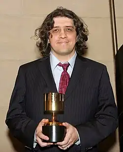 A man with short, curly brown hair wearing a black suit with a red tie holding an Annie Award, facing forward.