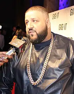 A man with short brown-blond hair and a dark beard speaks into a wireless microphone. He is wearing a shiny black shirt and has a dangling gold chain around his neck.