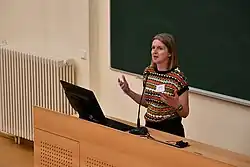 Dagmar Divjak teaching at the Summer School in Corpus Linguistics 2023, Tartu, Estonia. Divjak is standing behind a lectern with a computer on it and in front of a blackboard, and is gesturing animatedly.