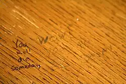 Written on a wooden desk, "You will die someday" a response is next to it.