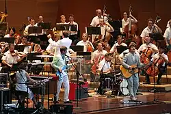 Five members of a band play keyboards, electric bass, drums and guitar in front of a symphony orchestra and its conductor, all dressed informally.