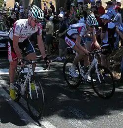 A pair of cyclists on the road, wearing matching white jerseys with blue and red trim. A car is partly visible behind them, and numerous spectators are on the roadside.