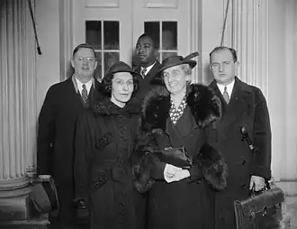 Five adults standing for a group photo: front is two white women; back row is two white men and a black man (center). They are indoors.