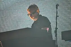 A color photograph of a man with short blonde hair on stage, wearing glasses and standing in front of a wall of white lights