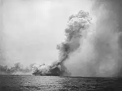 A black and white photograph showing a large cloud of smoke near the sea surface from which issues a towering mushroom cloud angled toward the right side of the photo