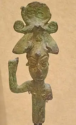 Photograph of an ancient bronze figurine with human features, raised arm, and ornate headdress. Worn texture suggests age.