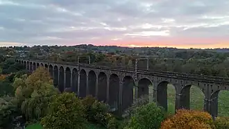 A photo of a landscape as viewed from a drone shot. It is a sunset in a valley with a arched railway viaduct cutting through the image. The viaduct has metal masts at periodic points and the bottom of the arches are partially covered in the photo by trees.