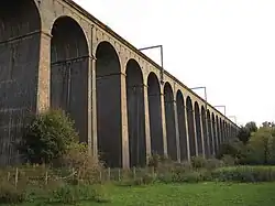 Part of a railway viaduct as viewed at an angle from up close on the ground. The brick arches of the viaduct are incredibly tall and there are periodic metal masts attached to the top, with some trees on the flat grassy ground.