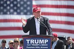 Trump speaking in front of an American flag behind a lectern, wearing a black business suit and a bright red "MAKE AMERICA GREAT AGAIN" baseball cap. The lectern sports a blue "TRUMP" sign.