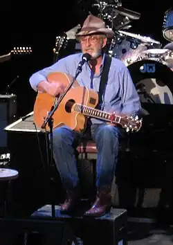 Don Williams, seated and playing an acoustic guitar while singing into a microphone