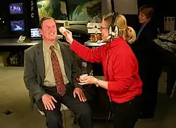 Man wearing a suit sitting in a chair in a studio getting his makeup touched up by a woman in a bright red sweater wearing headphones.