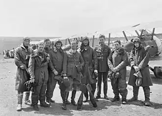 Nine men wearing a mixture of military uniforms with caps and flying suits with goggles, in front of a row of military biplanes