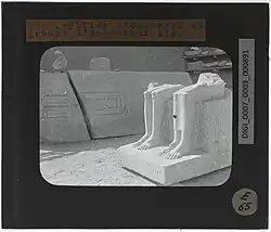 In the foreground, a broken statue of a seated couple, in the background, a large broken rectangular column inscribed with hieroglyphs