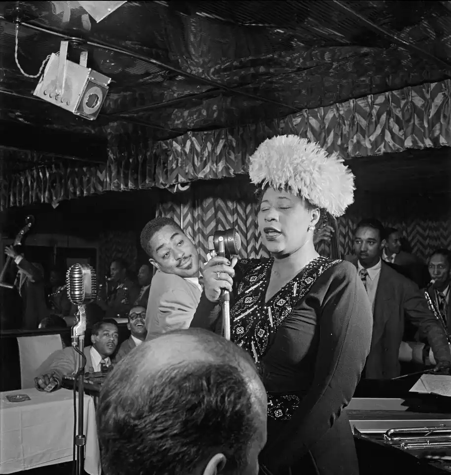 Fitzegeral is singing with a microphone inside a jazz club. Ray Brown stands behind her; Dizzy Gillespie and Milt Jackson sit in a table in the background; and Timme Roesenkratz in the foreground.