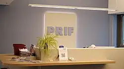 Modern reception desk with a plant, informational materials, and illuminated 'PRIF' sign on the wall.